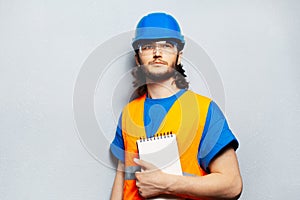 Portrait of young man, worker engineer wearing safety construction equipment, holding notebook against background of grey.