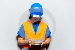 Portrait of young man, worker engineer wearing safety construction equipment, written in notebook against background of grey.