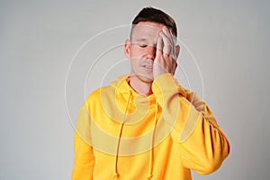 Portrait of young man in yellow hoodie with face palm gesture on gray background