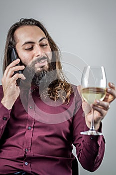 Portrait of a young Middle Eastern businessman with a beard and long hair holding a glass of white wine while having a phone conve photo