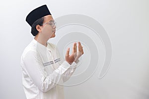 Portrait of young millennial Asian muslim man praying earnestly with his hands raised. Isolated image on white background