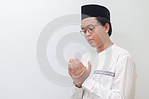 Portrait of young millennial Asian muslim man praying earnestly with his hands raised. Isolated image on white background