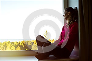 Portrait of young modern long-haired woman sitting on the windowsill with crossed legs and looking out at window.