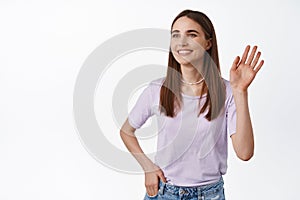 Portrait of young modern woman wave hello, looking aside at people and smiling, greeting, seeing friend and waving hi