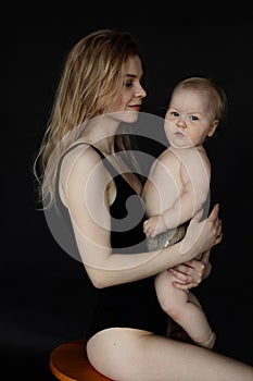Portrait of young mother in bodysuit sitting on chair and holding cute naked baby on black background. Happy parent