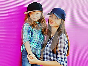 Portrait of young mother with child little girl wearing a checkered shirts and baseball cap in city over pink