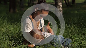 Portrait of young mother feeding baby in park, sitting on ground. Woman being playful with child while breastfeeding