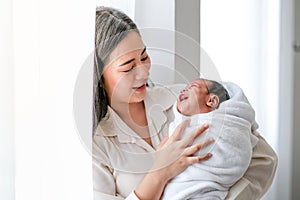 Portrait of young mother look at her newborn and they smile together in front of white curtain with morning light