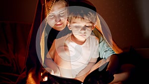 Portrait of young mother lying with her little son under blanket and reading him story book