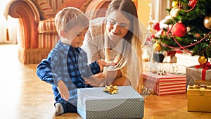 Portrait of young mother sitting with toddler boy under Christmas tree and looking at gift box with golden ribbon bow