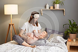 Portrait of young mother in sleep mask spending time with her baby daughter child, embracing and hugging kid while sitting on the