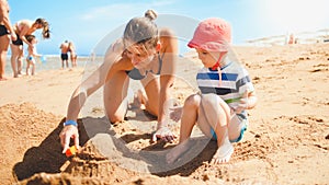 Portrait of young mother teaching her 3 years old toddler son building castles from sand on the sea beach. Family