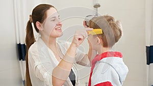 Portrait of young mother using hairdryer to dry hair of her little son after washing in bathroom. Concept of child