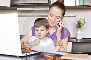 Portrait of young mother works freelance on laptop computer, communicates with someone via smart phone, looks after her little son