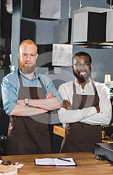 portrait of young multicultural owners of coffee shop in aprons standing with crossed arms