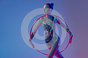 Portrait of young, muscular girl, female rhytmic gymnast training with hoop isolated over blue studio background in neon