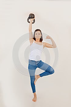 Portrait of young muscular woman wearing white top, blue jeans, standing on leg, lifting kettle bell, showing muscles.