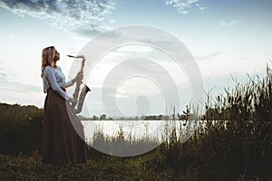 Portrait of young musician on nature background, woman playing saxophone on bank river in reeds, concept music and relax