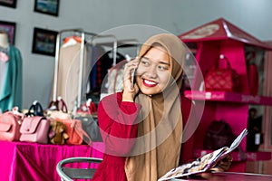 Portrait of a young muslim entrepeneur sitting and smile talking in her smartphone in front of fashion boutique as background -