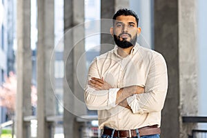 Portrait of a young Muslim man in a shirt standing outside an office building, crossing his arms over his chest and