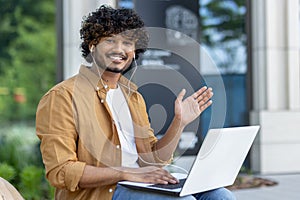 Portrait of a young Muslim man wearing headphones, holding a laptop on his lap, sitting on a bench in a city street