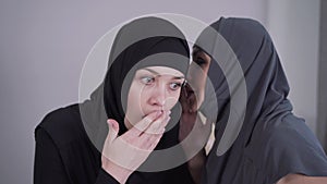 Portrait of young Muslim woman in hijab listening to friend whispering on her ear. Surprised woman covering mouth with