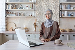 Portrait of young Muslim woman in hijab, woman in glasses looking at camera studying at home sitting in kitchen, female
