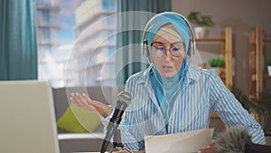 Portrait young muslim woman records a podcast while sitting in front of a microphone in her living room