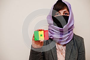 Portrait of young muslim woman wearing formal wear, protect face mask and hijab head scarf, hold Senegal flag card against photo