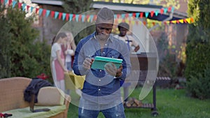 Portrait of young overburdened African American man sighing holding tablet with relaxed friends cooking bbq at