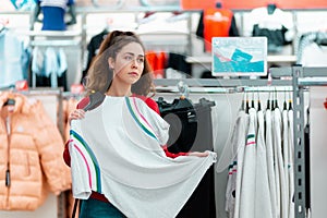 Portrait of a young pensive woman choosing white t-shirt in a store. The concept of shopping and consumerism