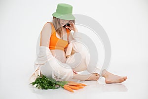 Portrait of young perfect pregnant woman with long dark hair wearing green bucket hat, sitting near bunch of carrots.