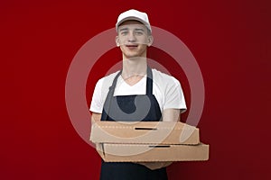 Portrait of a young pizza delivery man in uniform on a red background, a courier guy gives boxes of pizza