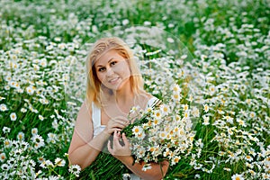 Portrait of a young, plump, beautiful woman resting on a chamomile field at sunset. A plus-size woman in a white sundress sits