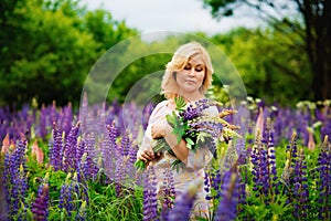 Portrait of a young plump blonde woman in a blooming field of lupines. A woman collects a bouquet of lilac-pink flowers in a