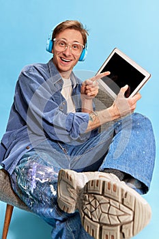 Portrait of young positive man in headphones sitting on chair and poiting on tablet over blue studio background. News