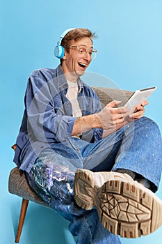 Portrait of young positive man sitting on chair in headphones and watching movie on tablet over blue studio background