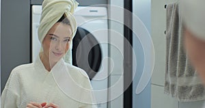Portrait of young positive woman wearing bathrobe and towel on head after shower smiling to camera with mirror