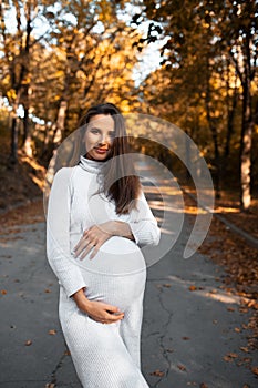 Portrait of young pregnant smiling woman with hands on belly, wearing white dress on background of golden autumn park.
