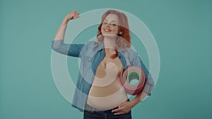 Portrait of a young pregnant woman holding a yoga mat in the studio on a blue background, showing a strong gesture. A