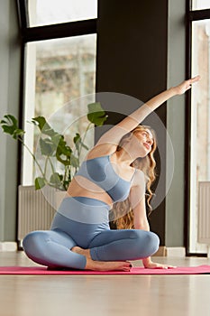 Portrait of young pregnant woman in sportswear doing yoga, sitting on yoga mat and doing stretching indoors on a daytime