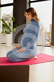 Portrait of young pregnant woman in sportswear sitting on yoga mat and meditating. Stretching exercises