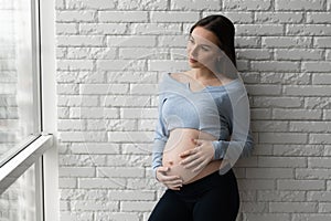 Portrait of a young pregnant woman. Stands against a brick wall and looks out the window.