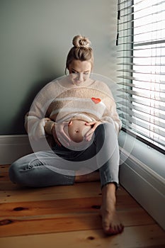 Portrait of young pregnant woman wearing beige sweater with application of heart, sitting on floor stroking naked belly.