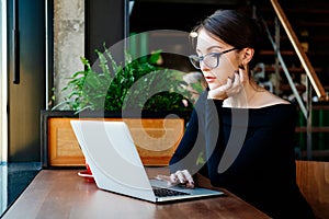 Portrait of young pretty business woman in glasses sitting on cafe and using laptop