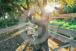 Portrait of a young pretty caucasian girl sitting on a park bench taking a selfie at sunset