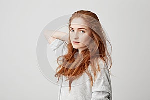 Portrait of young pretty ginger girl with freckles looking at camera touching hair over white background.