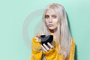 Portrait of young pretty girl with blue eyes, holding black cup on background of pastel green of color. Wearing yellow sweater.