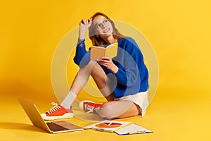 Portrait of young pretty girl, student wearing eyeglasses sitting on floor surrounding books and laptop over yellow