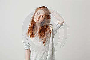 Portrait of young pretty redhead girl with freckles looking at camera smiling touching hair over white background.